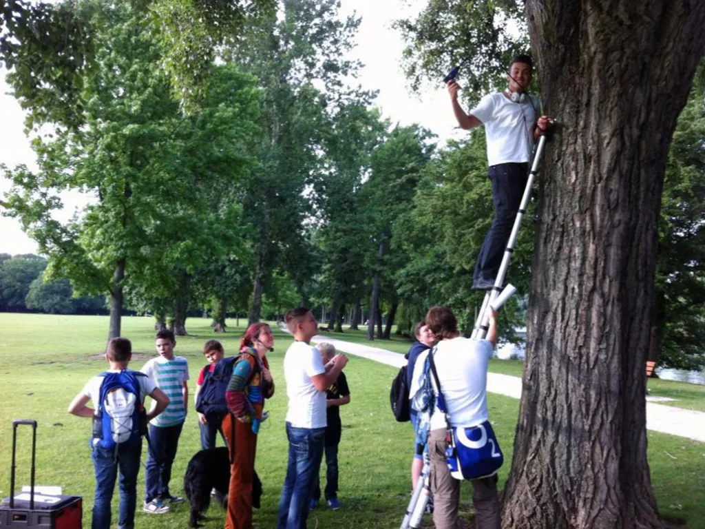 Schülergruppe steht unter einem großen Baum, eine Person klettert an einer Leiter am Stamm hinauf.